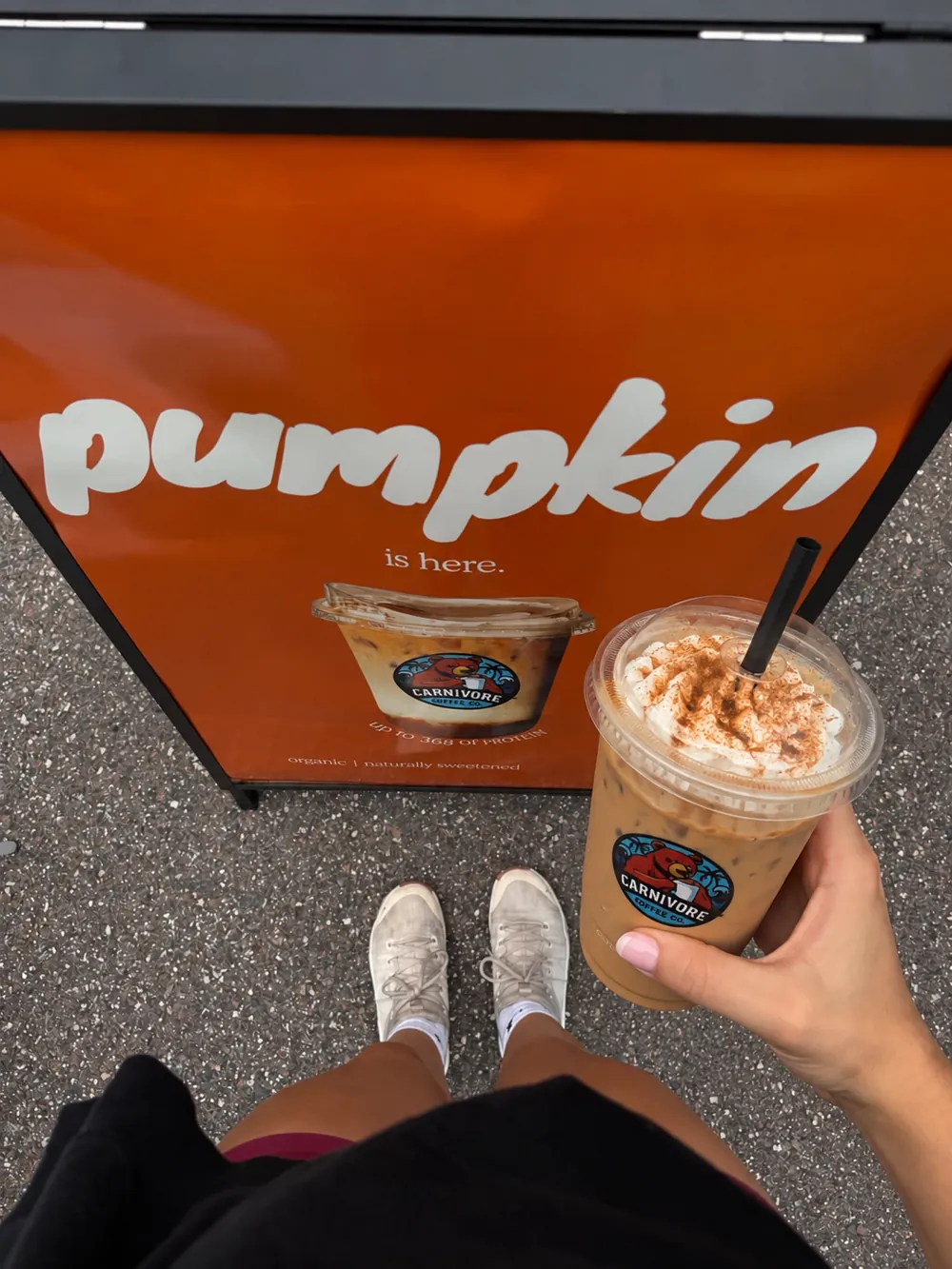 Carnivore Coffee pumpkin iced latte held up in front of the pumpkin is here sidewalk sign